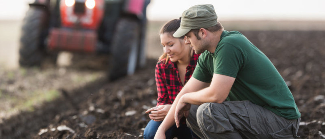 Deux jeunes agriculteurs regardent la terre dans un champ, avec un tracteur en fond.